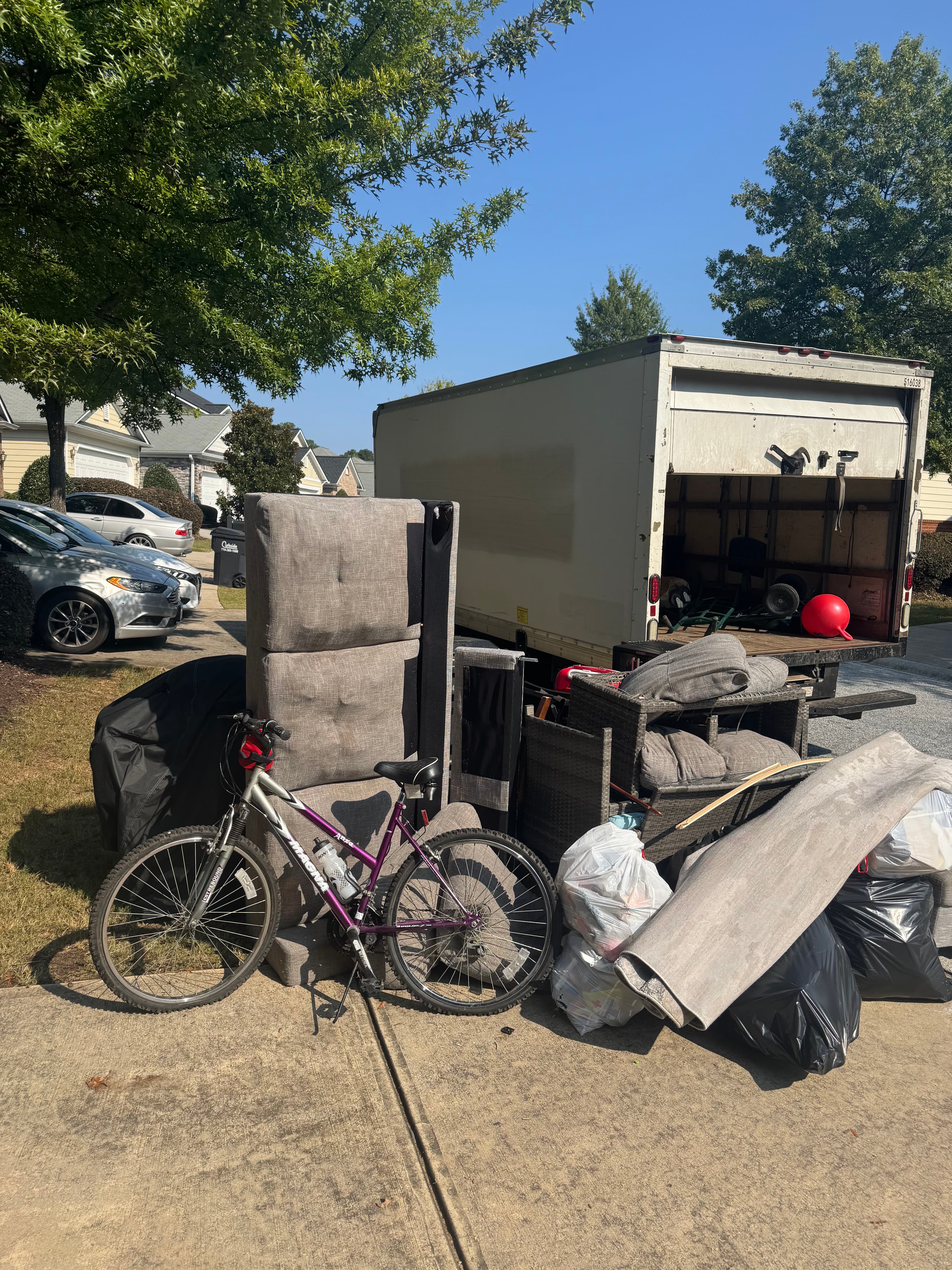 Junk removal truck backed into a driveway while furniture, bags, and bulky items are staged for loading.